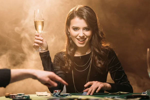 Attrayant fille heureuse tenant un verre de champagne à la table de poker dans le casino et regardant la caméra — Photo de stock