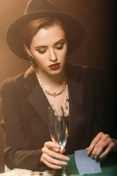 Chica atractiva en chaqueta y sombrero sosteniendo copa de champán y jugando al póquer en el casino - foto de stock