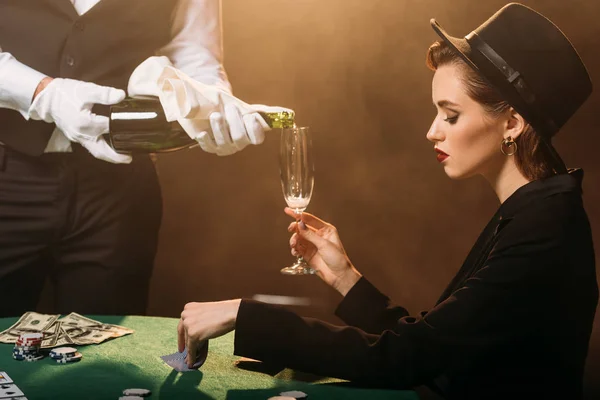 Waiter pouring champagne in glass of attractive girl in jacket and hat while she playing poker in casino — Stock Photo