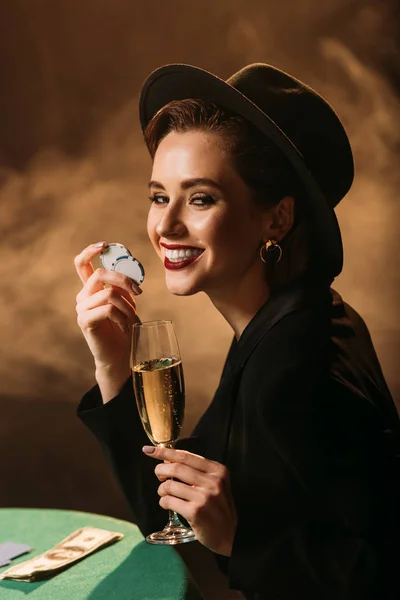 Chica atractiva feliz en chaqueta y sombrero sosteniendo la copa de champán y fichas de póquer en la mesa en el casino, mirando a la cámara - foto de stock