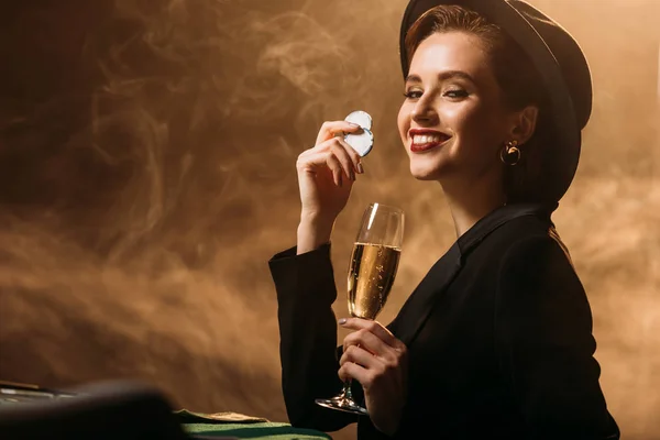 Chica atractiva sonriente en chaqueta y sombrero sosteniendo copa de champán y fichas de póquer en la mesa en el casino - foto de stock