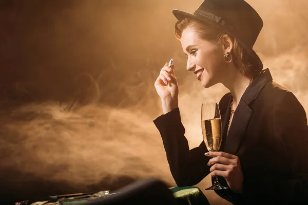 Вид сбоку улыбающейся привлекательной девушки в куртке и шляпе с бокалом шампанского и фишками для покера за столом в казино — стоковое фото