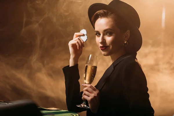 Attractive girl in jacket and hat holding glass of champagne and poker chips at table in casino — Stock Photo