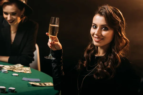 Smiling attractive girl holding glass of champagne at poker table in casino and looking at camera — Stock Photo