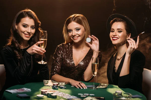 Garotas atraentes sorridentes com copo de champanhe, cigarro e fichas de poker sentadas à mesa no cassino — Fotografia de Stock