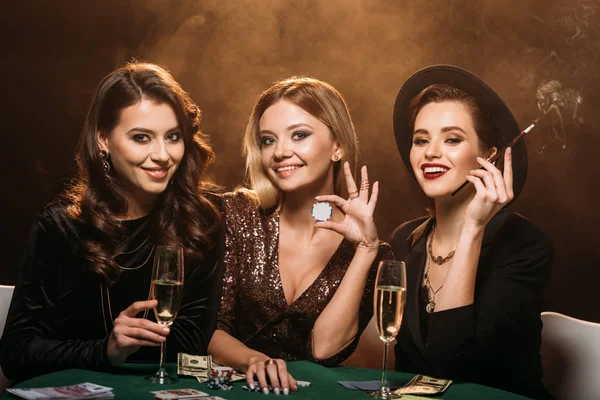 Felices amigos atractivos con copa de champán, cigarrillos y fichas de póquer sentado en la mesa en el casino y mirando a la cámara - foto de stock