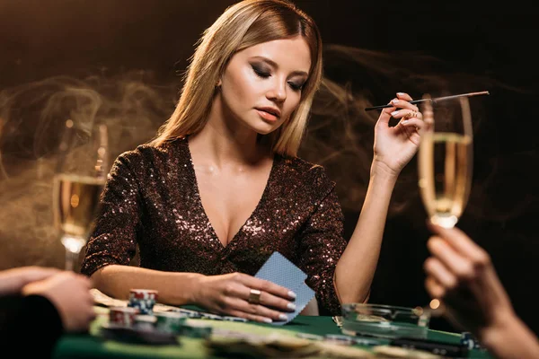 Menina atraente segurando cigarro e cartas de poker na mesa no cassino — Fotografia de Stock