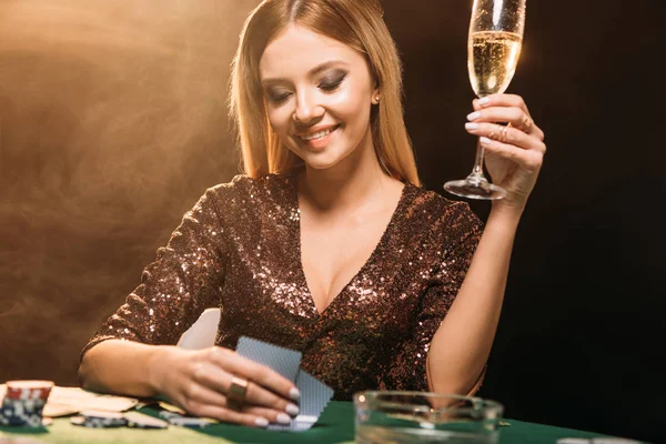 Smiling attractive girl holding glass of champagne and looking at poker cards at table in casino — Stock Photo