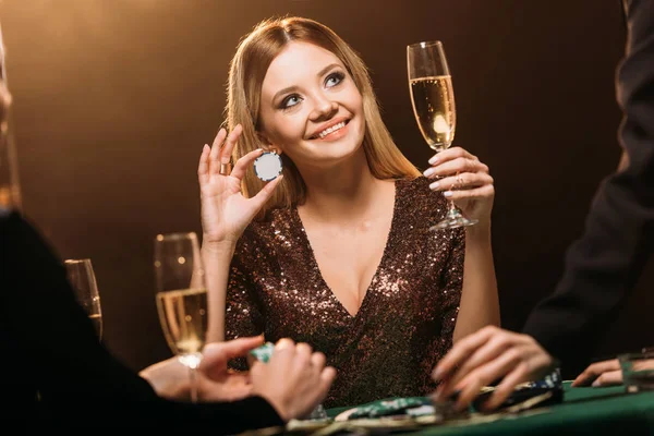 Beautiful happy woman holding glass of champagne and poker chip at table in casino and looking up — Stock Photo