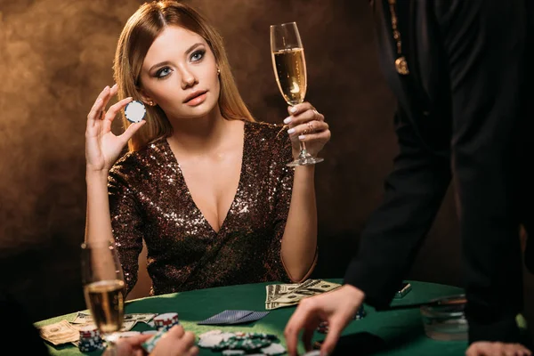 Beautiful woman holding glass of champagne and poker chip at table in casino — Stock Photo