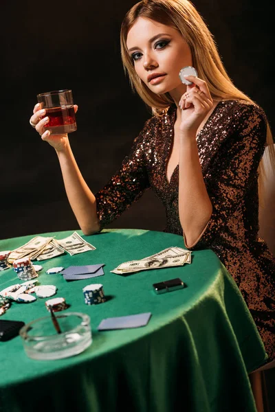 Atractiva chica sonriente sosteniendo vaso de whisky y fichas de póquer en la mesa en el casino, mirando a la cámara - foto de stock