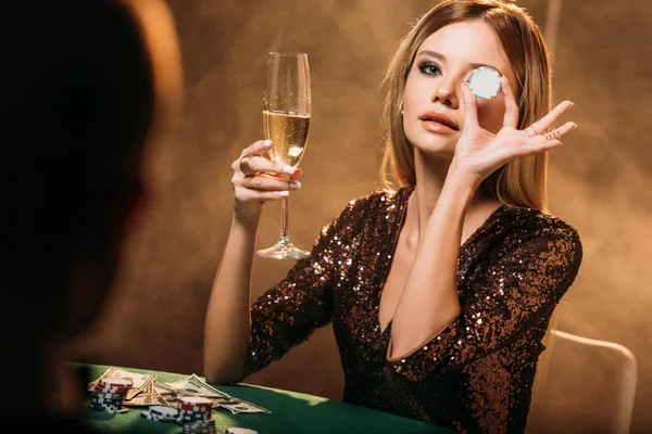 Menina atraente segurando copo de champanhe e olho de cobertura com chip de poker na mesa no cassino — Fotografia de Stock