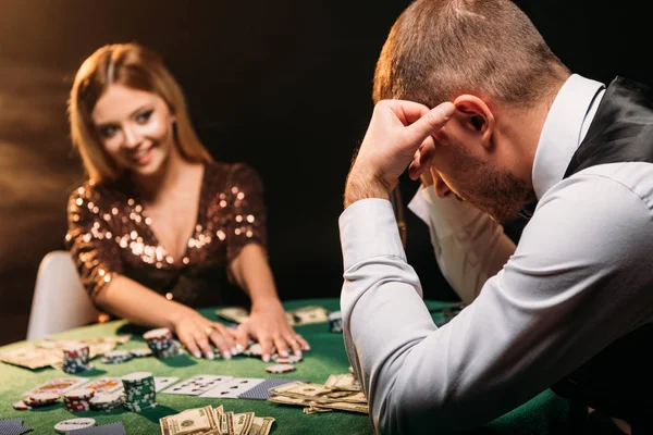 Улыбающаяся привлекательная девушка берет фишки для покера и смотрит на грустного крупье в казино — стоковое фото
