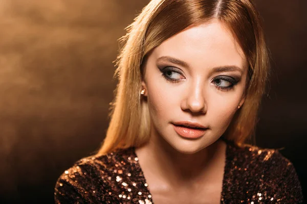 Retrato de chica atractiva con maquillaje en vestido de fiesta brillante mirando hacia otro lado en marrón - foto de stock