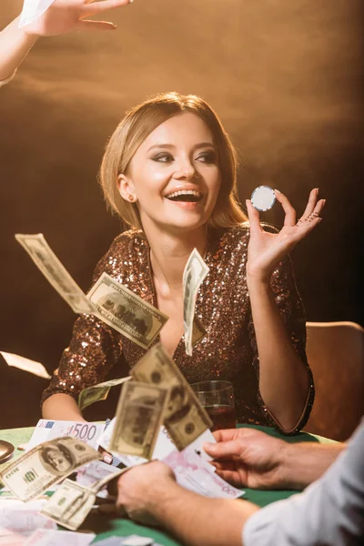 Excited attractive girl holding poker chip with croupier at casino, dollar banknotes falling on table — Stock Photo