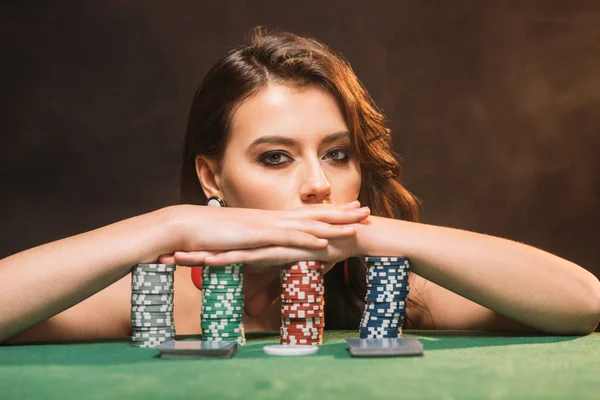 Beautiful brown haired girl leaning on poker chips and looking at camera isolated on black — Stock Photo