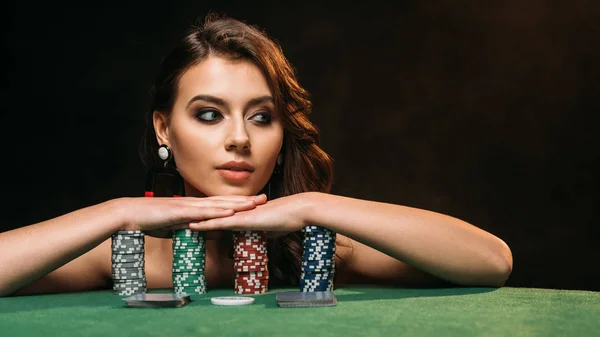 Beautiful brown haired girl leaning on poker chips and looking away isolated on black — Stock Photo