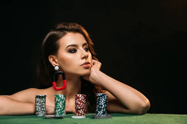Bela menina de cabelos castanhos descansando queixo na mão e olhando para longe, fichas de poker na mesa isolada em preto — Fotografia de Stock