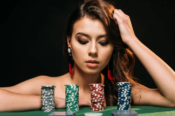 Bela menina de cabelos castanhos inclinando-se na mesa e olhando para fichas de poker isolado em preto — Fotografia de Stock