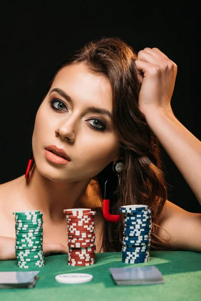Hermosa chica de cabello castaño apoyado en la mesa con fichas de póquer y mirando a la cámara aislada en negro - foto de stock