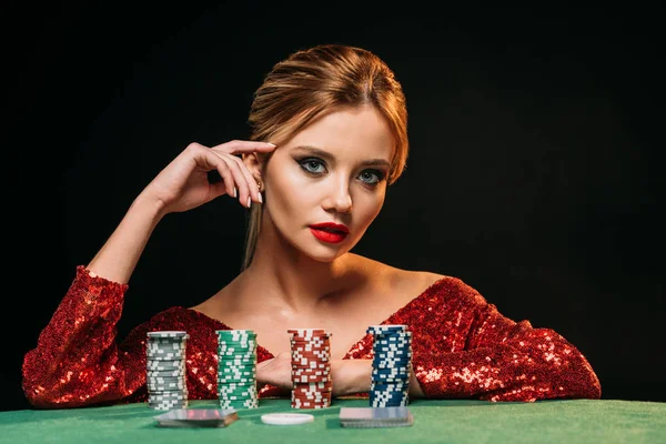 Attractive girl in red shiny dress leaning on table with poker chips and looking at camera isolated on black — Stock Photo