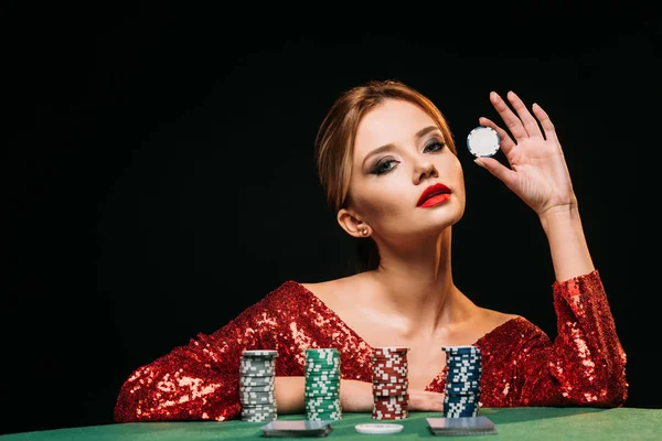 Menina atraente em vestido vermelho brilhante inclinando-se na mesa, segurando chip de poker e olhando para a câmera isolada no preto — Fotografia de Stock