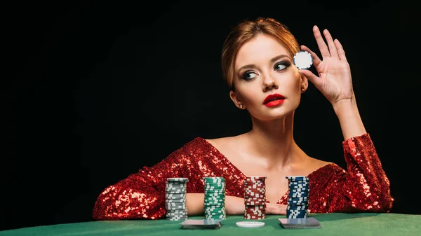 Attractive girl in red shiny dress leaning on table, holding poker chip and looking away isolated on black — Stock Photo