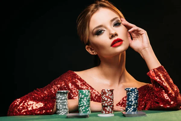 Beautiful girl in red shiny dress leaning on table with poker chips and looking at camera isolated on black — Stock Photo