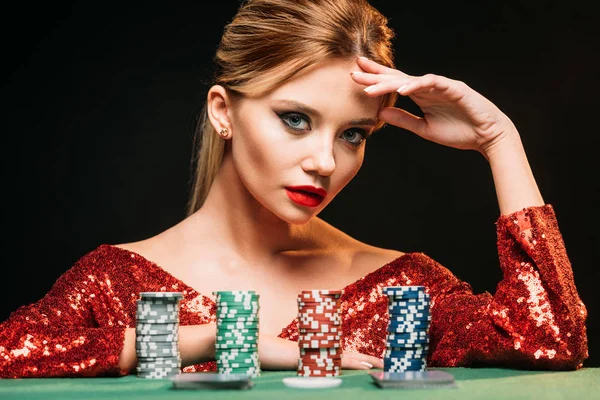 Attraente ragazza in abito rosso scintillante appoggiato sul tavolo con chip di poker isolato su nero — Foto stock