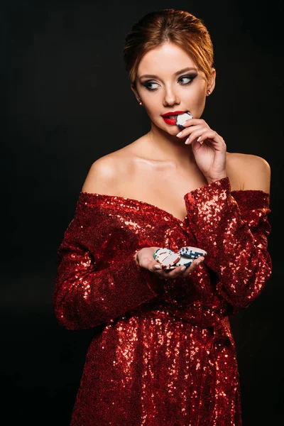 Beautiful girl in red shiny dress holding and biting poker chips isolated on black — Stock Photo