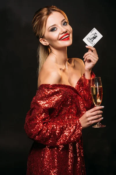 Lächelndes attraktives Mädchen in rotem, glänzendem Kleid mit Jokerkarte und einem Glas Champagner auf schwarzem Grund, das in die Kamera blickt — Stockfoto