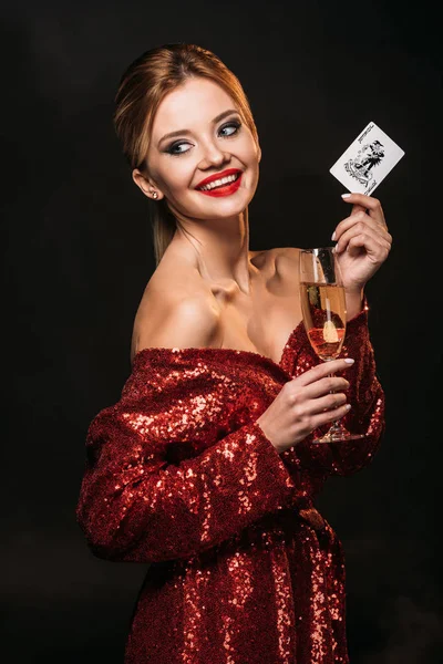 Smiling attractive girl in red shiny dress holding joker card and glass of champagne isolated on black, looking away — Stock Photo