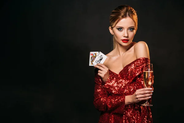 Attractive girl in red shiny dress holding joker and queen of hearts cards isolated on black, looking at camera — Stock Photo