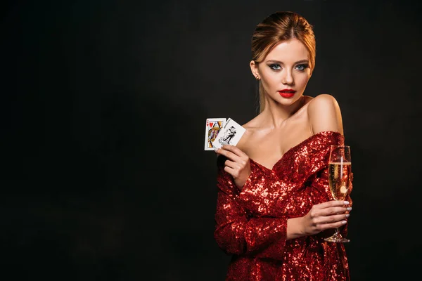 Attrayant fille en robe rouge brillant tenant joker et reine des cartes de coeur isolé sur noir, regardant caméra — Photo de stock