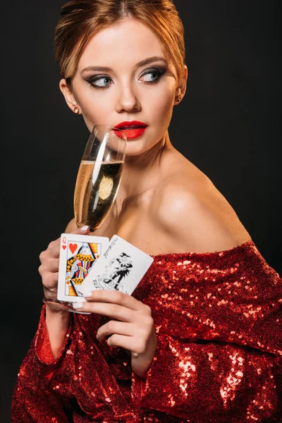 Jolie fille en robe rouge brillant tenant joker et reine des cartes de coeur, boire du champagne isolé sur noir — Photo de stock