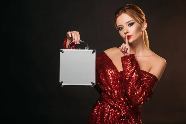 Hermosa mujer en rojo brillante vestido sosteniendo caja de dinero y mostrando gesto de silencio aislado en negro - foto de stock