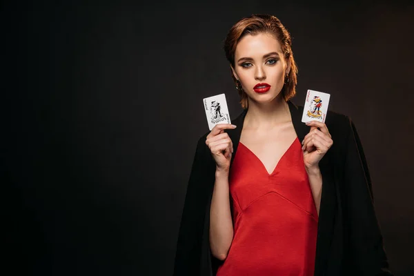 Beautiful girl in red dress and black jacket holding poker cards and looking at camera isolated on black — Stock Photo