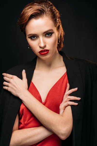 Chica atractiva en vestido rojo y chaqueta negra de pie con los brazos cruzados y mirando a la cámara aislada en negro - foto de stock