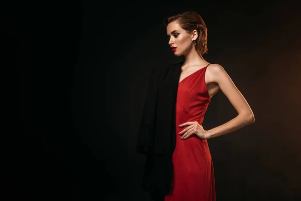 Приваблива дівчина в червоній сукні і чорна куртка, що стоїть ізольовано на чорному — стокове фото