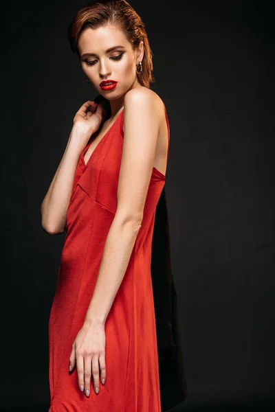 Chica atractiva en vestido rojo sosteniendo chaqueta negra y mirando hacia abajo aislado en negro - foto de stock