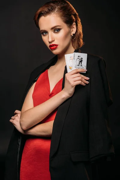 Chica atractiva en vestido rojo y chaqueta negra con tarjetas de póquer y mirando a la cámara aislada en negro - foto de stock