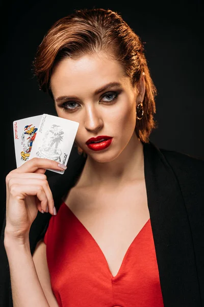 Портрет привабливої дівчини в червоній сукні і чорній куртці, що тримає покерні карти і дивиться на камеру ізольовано на чорному — стокове фото