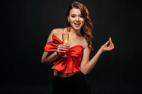 Excitada chica de cabello castaño en corsé rojo sosteniendo copa de champán y chips de casino aislados en negro - foto de stock