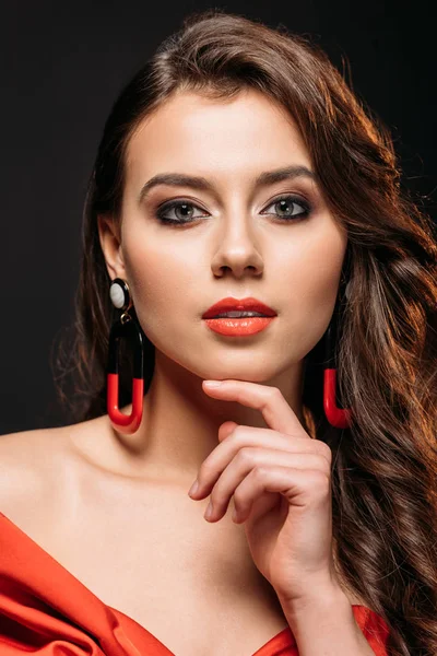 Portrait of beautiful brown haired girl in red corset and earrings touching chin isolated on black — Stock Photo