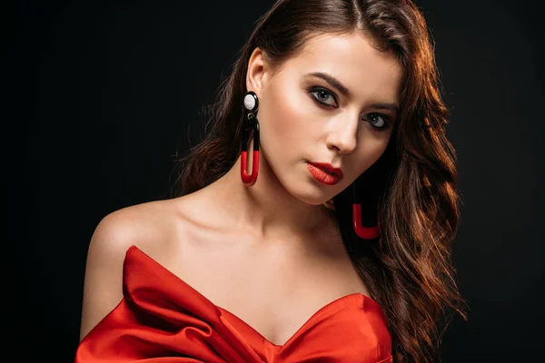 Portrait of beautiful brown haired girl in red corset and earrings looking at camera isolated on black — Stock Photo
