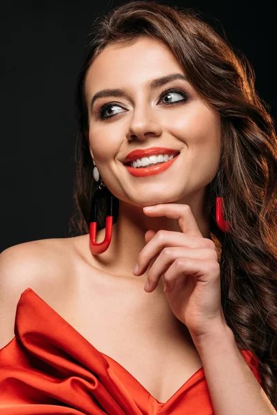 Portrait de sourire belle fille aux cheveux bruns en corset rouge et boucles d'oreilles regardant loin isolé sur noir — Photo de stock