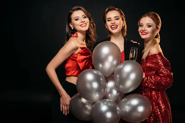 Smiling attractive girls in stylish party clothes holding bundle of grey balloons and looking at camera isolated on black — Stock Photo