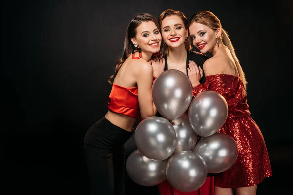 Улыбающиеся привлекательные девушки в стильной одежде для вечеринок, держащие связку серых воздушных шаров, обнимающие и смотрящие на камеру, изолированную на черном — стоковое фото