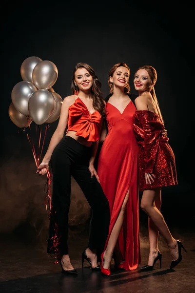Smiling attractive girls in fashionable party clothes holding bundle of grey balloons and looking at camera on black — Stock Photo