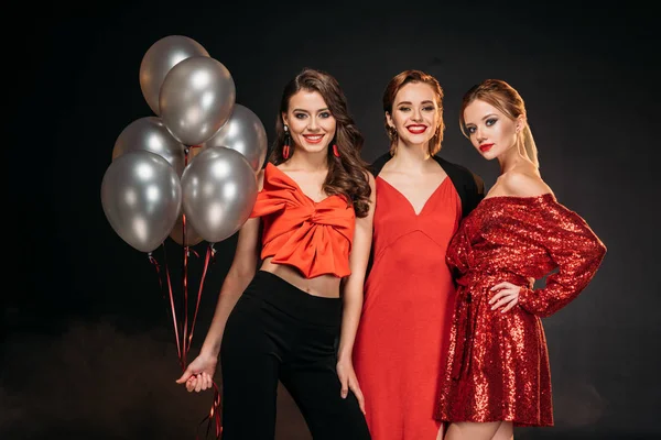 Sorrindo meninas atraentes em roupas vermelhas segurando pacote de balões cinza isolado em preto — Fotografia de Stock